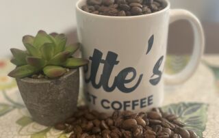 Everette’s Fresh Roast Coffee mug filled with roasted coffee beans next to a small succulent plant on a floral table.