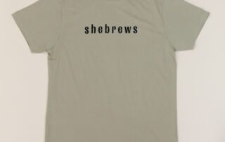 Front of sage green She Brews t-shirt with minimalist black “shebrews” text
