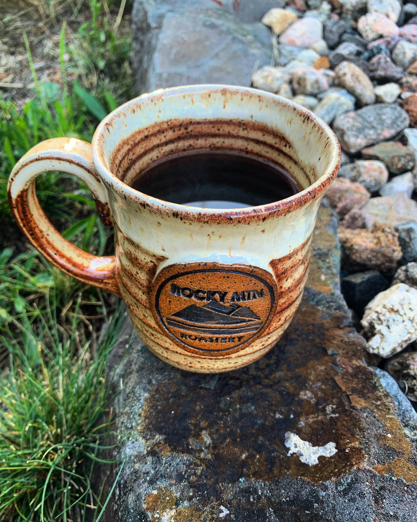 Rustic Rocky Mountain Roastery coffee mug filled with coffee sitting on a rock outdoors