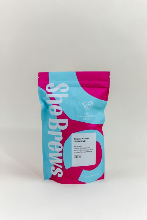 Front view of She Brews Rwanda Natural Single Origin Coffee 12oz bag featuring vibrant pink and blue design with flavor notes of brown sugar, dried cherries, hibiscus, and lime.